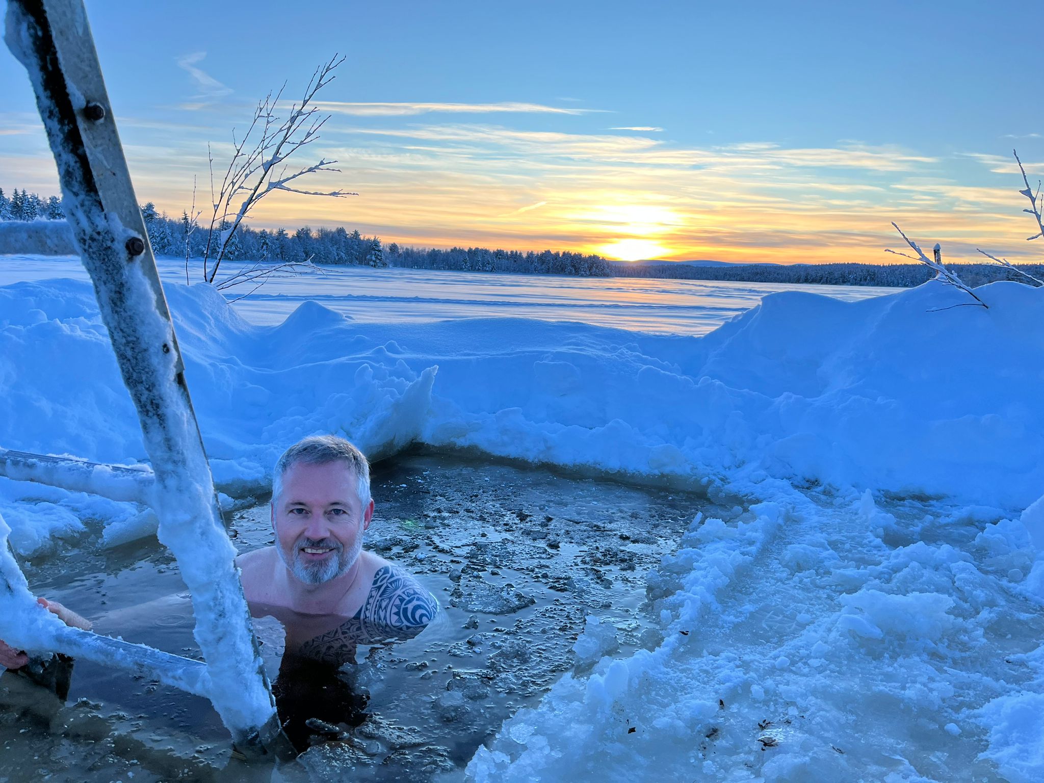 Un homme à la barbe grise et aux tatouages dans un bain de glace dans un trou creusé dans un lac gelé, entouré de neige, au coucher du soleil